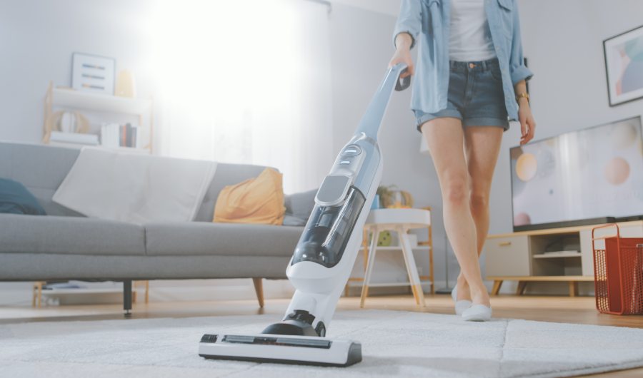 Primer Plano De Una Mujer Joven Y Guapa Con Camisa Vaquera Y Pantalones Cortos Aspirando Una Alfombra En Una Habitación Luminosa Y Acogedora En Casa. Utiliza una moderna aspiradora sin cable. Está Feliz y Alegre.